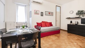 A compact living space with a dining area, a bed with red bedding, a small kitchenette, and a wall-mounted clock. - Easylife - Modern apartment in Bocconi (Milano)