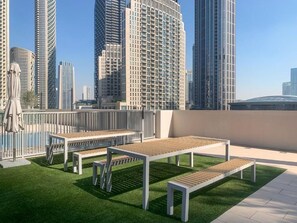 A rooftop terrace with white benches and artificial grass, surrounded by high-rise buildings. - Burj Khalifa & Dubai Mall Proximity : London Condo (Dubai)