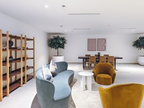 A modern living room with a sofa, armchairs, a coffee table, a wooden bookshelf, a dining area with chairs, and two potted plants. - Burj Khalifa & Dubai Mall Proximity : London Condo (Dubai)