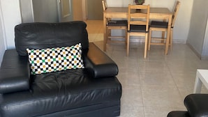 Una sala de estar con un sofá de cuero negro, un cojín a cuadros, una mesa de comedor de madera con sillas y una pequeña planta en maceta.