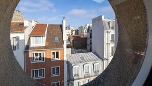 Une vue à travers une fenêtre circulaire sur un paysage urbain composé de différents bâtiments, dont un imposant bâtiment en briques rouges et un bâtiment blanc avec un toit gris.