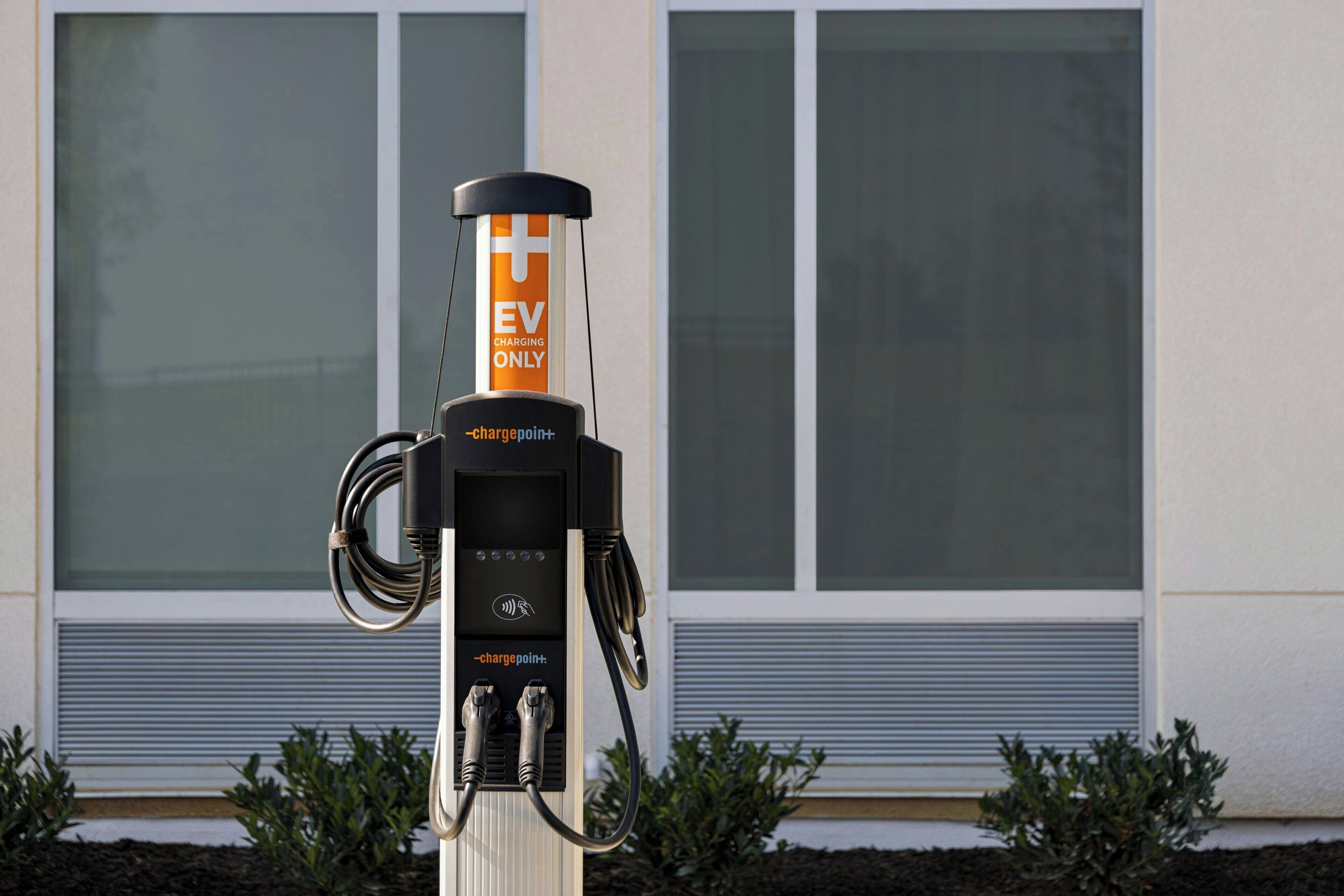Een laadstation voor elektrische voertuigen met een label dat aangeeft dat het uitsluitend voor EV-opladen is, verbonden met een gebouw met grote ramen en een metalen hekwerk.