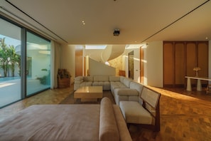 Une chambre d’hôtel moderne avec un grand lit, un canapé d’angle, une petite table et un banc. Une grande fenêtre offre une vue sur les palmiers et une piscine.