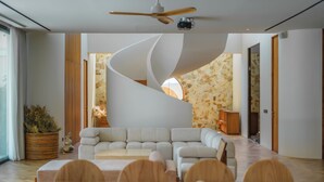 A modern living room with a curved white wall, a sectional sofa, wooden flooring, and a dining area with a table and chairs. - Babylon (Cemagi)