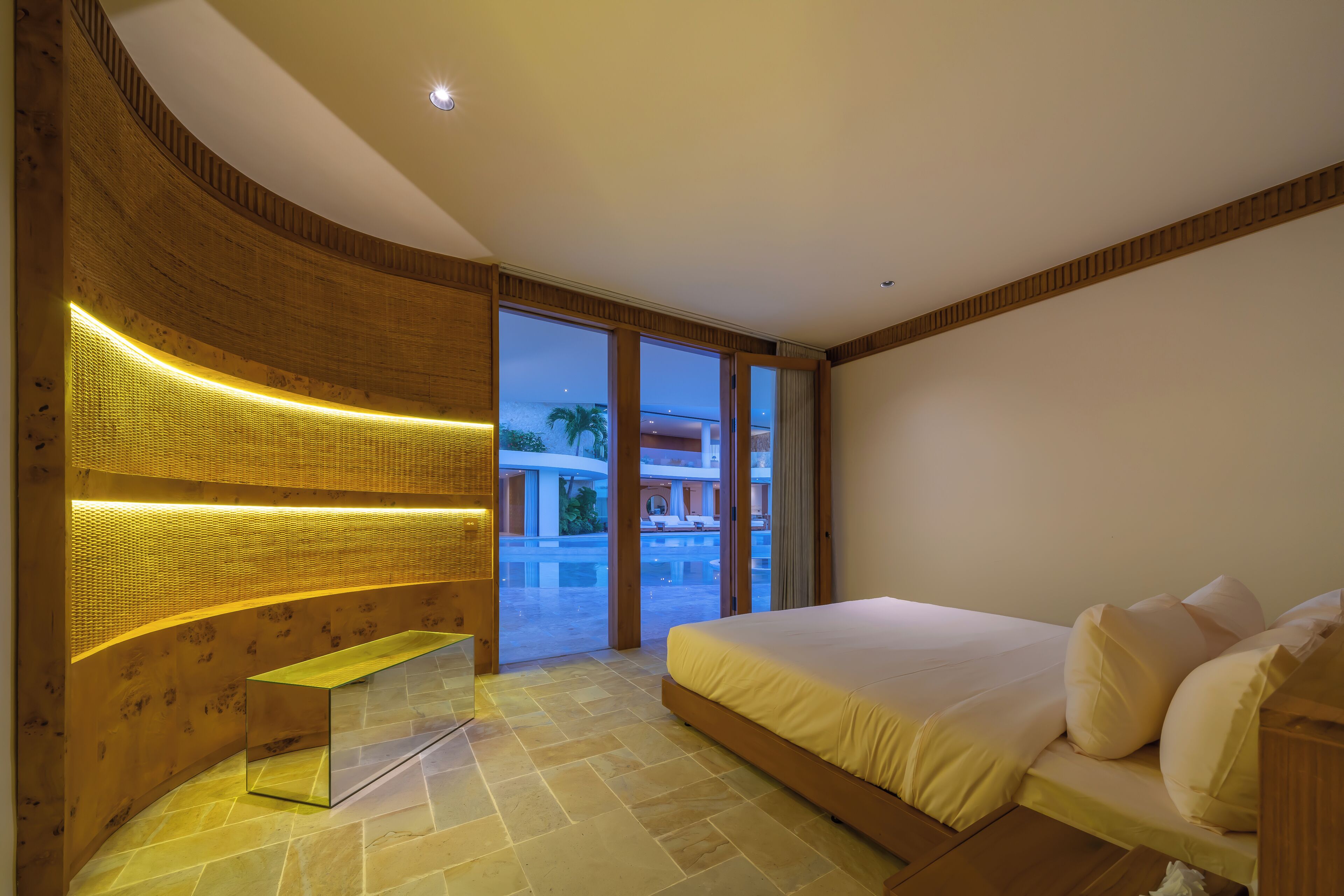 Une chambre moderne avec un grand lit, un banc intégré et une vue sur l’espace extérieur abritant une piscine.