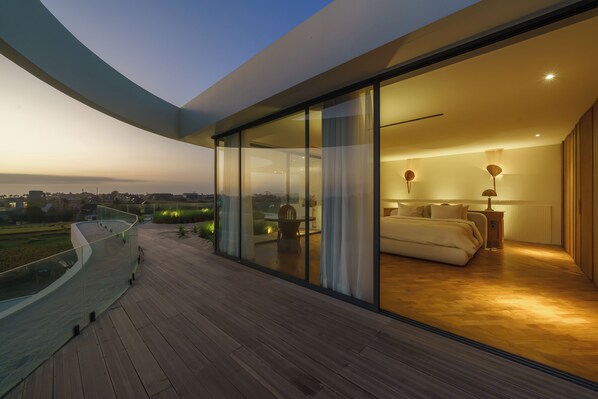 A modern bedroom with a glass wall, a curved balcony, and a view of the cityscape. - Babylon (Cemagi)