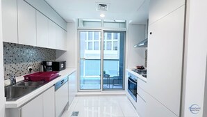 A modern kitchen with white cabinets, a tiled backsplash, a microwave, a red tray, and a view to a balcony with a stove and a chair. - Chic One Bedroom Near Dubai Mall in Damac Voleo (Business Bay)