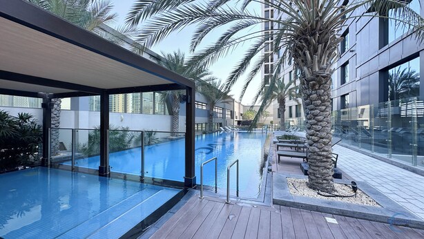 A modern indoor swimming pool with clear glass walls, surrounded by palm trees and lounge chairs. - Chic One Bedroom Near Dubai Mall in Damac Voleo (Business Bay)