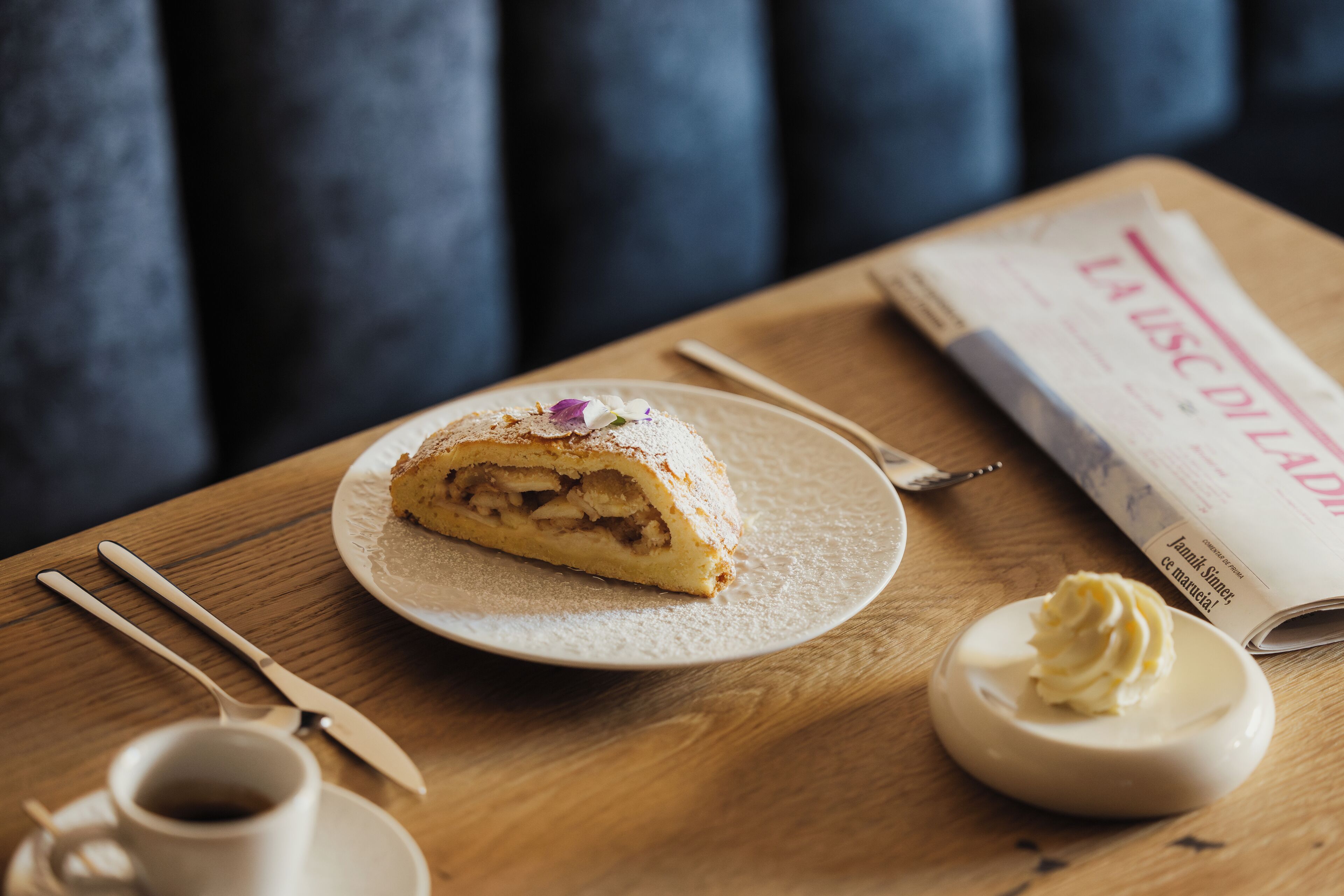 Una rebanada de tarta de manzana en un plato, con una cuchara y una taza de café sobre una mesa de madera.