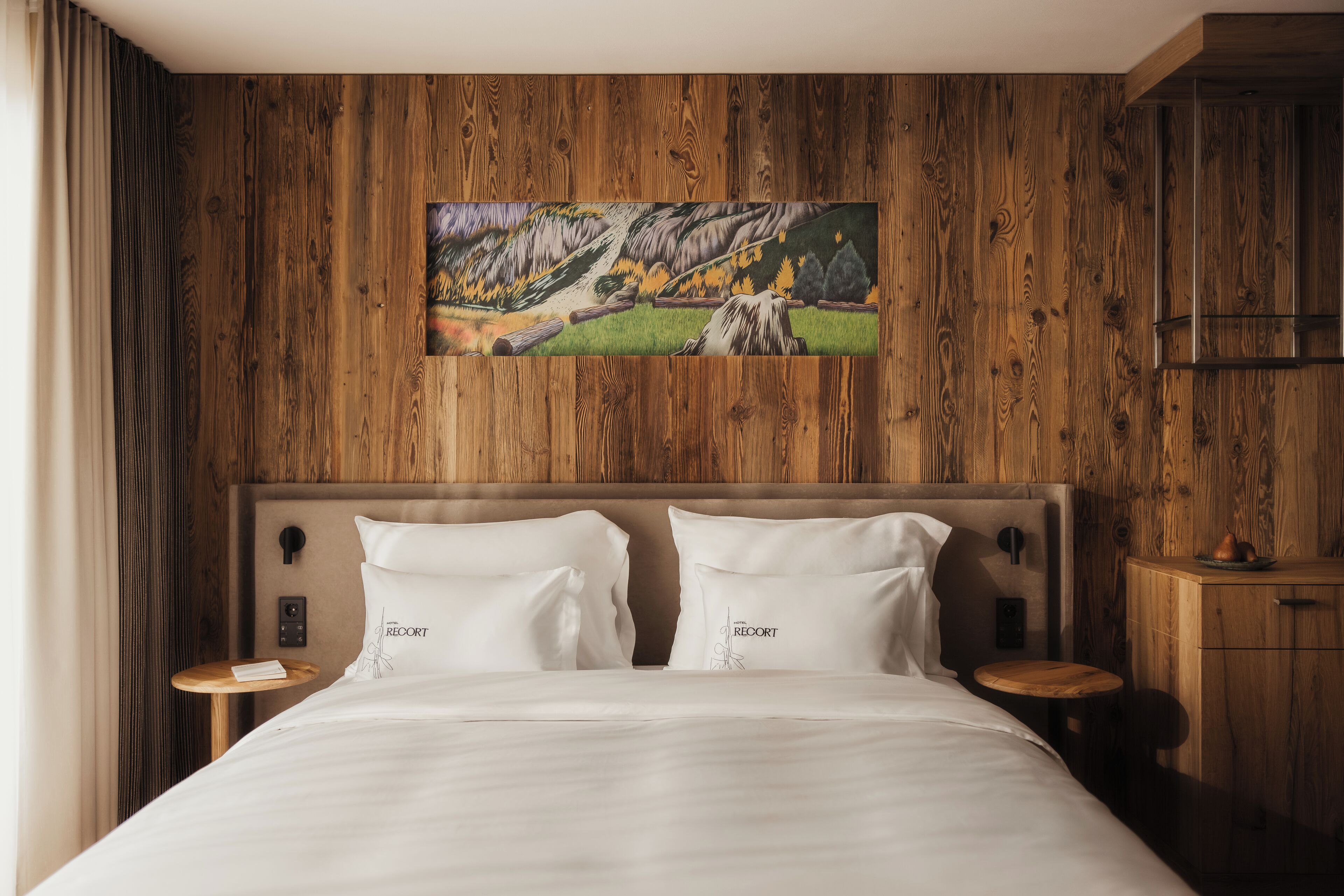 A hotel room with a wooden wall, a painting of a landscape, a bed with two pillows, and a nightstand with a lamp.
