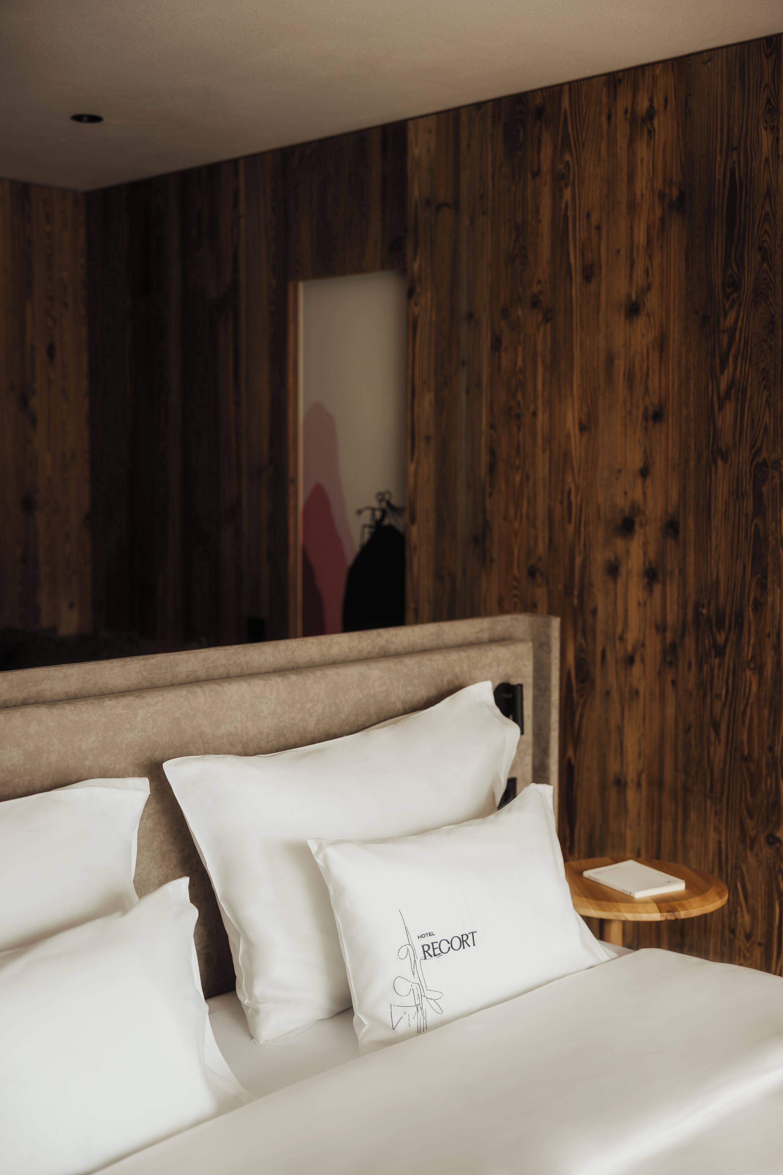 Una cama bien hecha con sábanas y almohadas blancas, un cabecero de madera y una mesita de noche de madera con un libro.