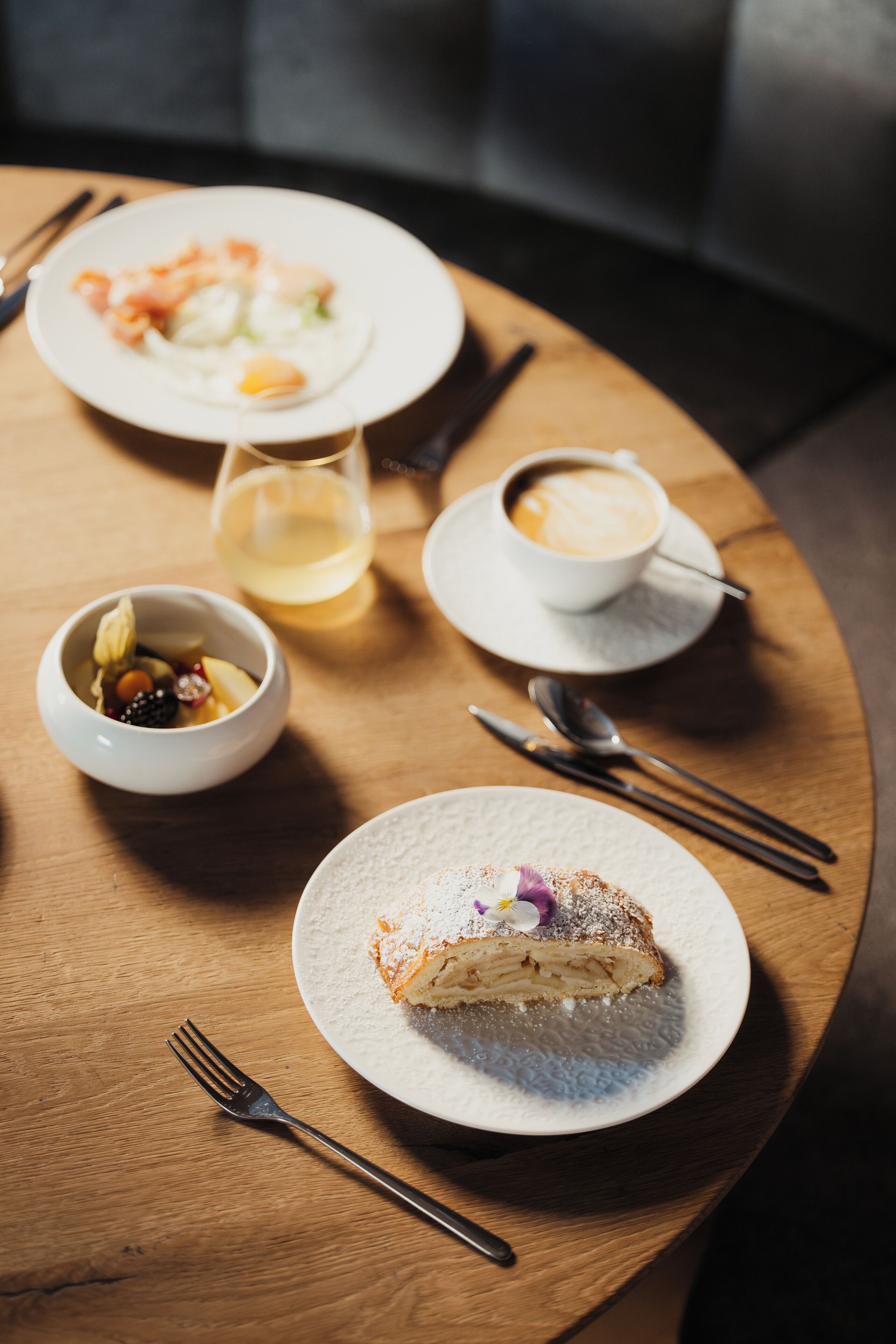 A wooden table with a dessert plate, a bowl of fruit, a cup of coffee, and a glass of white wine.