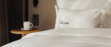A hotel room with a wooden headboard, a painting of a rabbit, a white pillow with "RECORD" text, a bedside table with a white cup, and a power outlet.