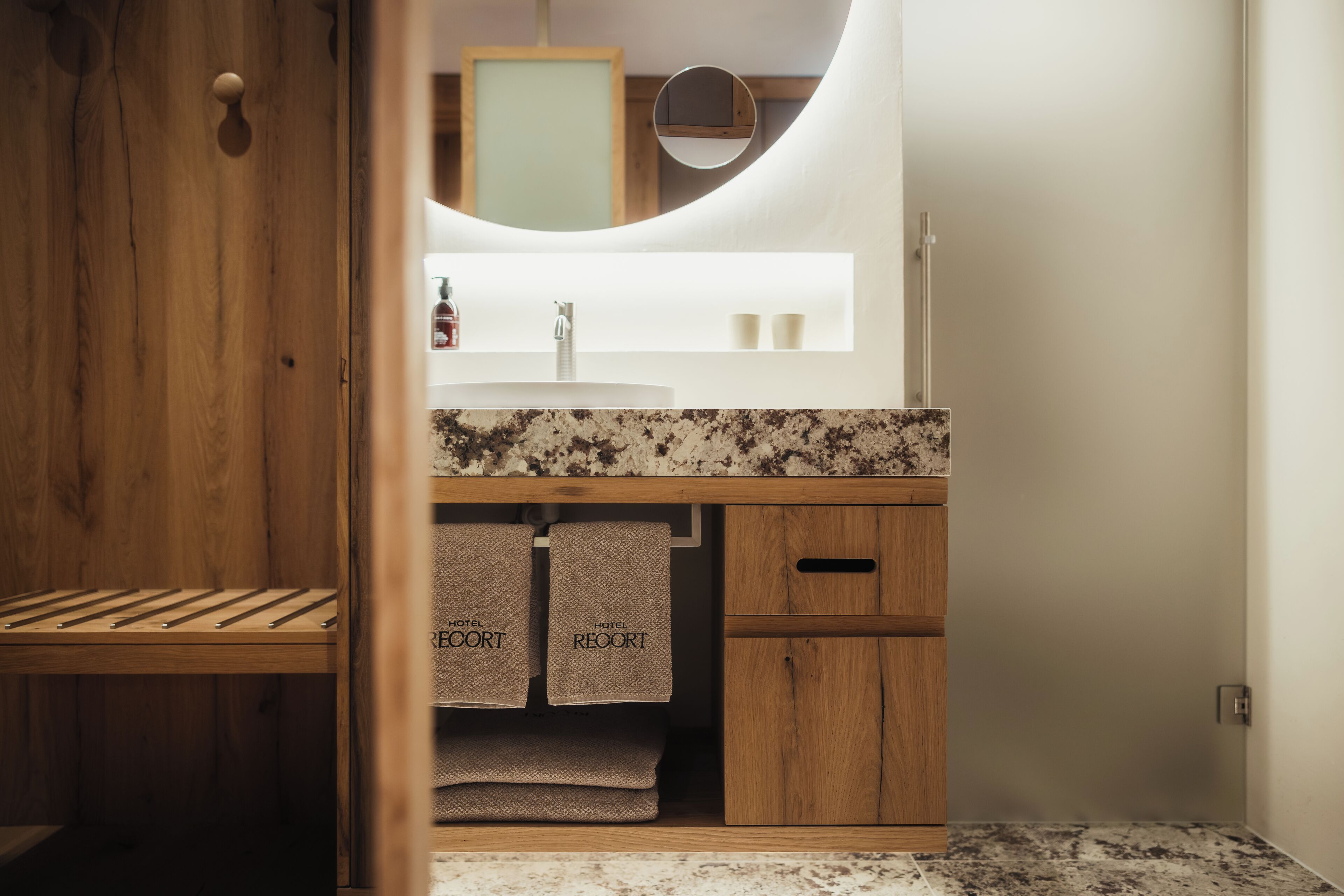 Un banc en bois, un lavabo avec un plan de travail en marbre, un miroir et des serviettes étiquetées « HÔTEL ».