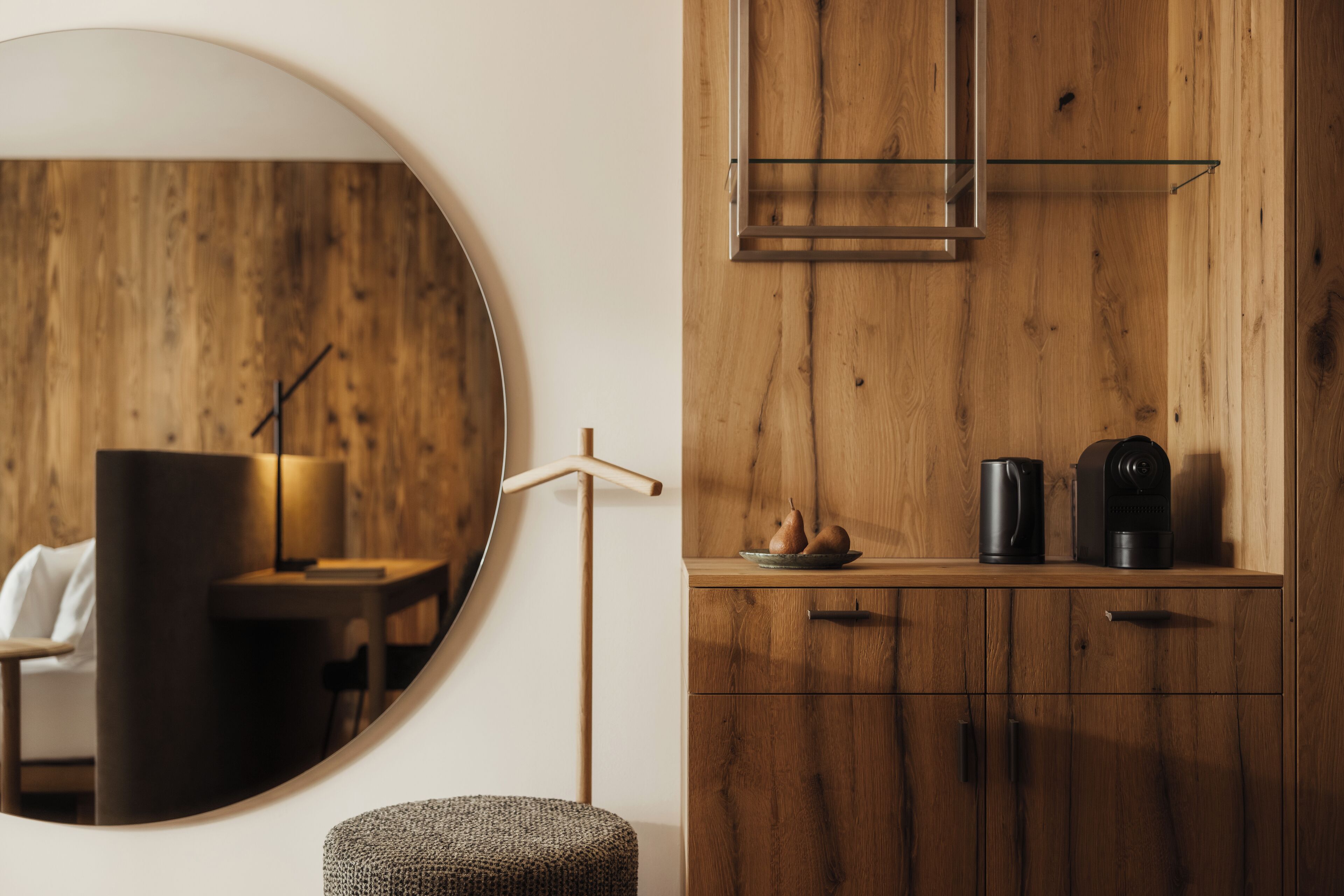 A modern room with a round mirror, a wooden cabinet with a glass shelf, and a small round ottoman.