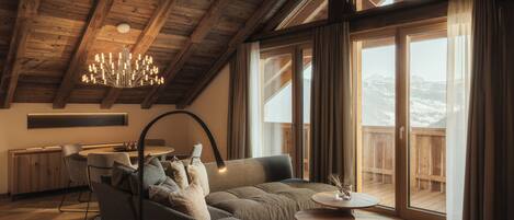 A spacious living area with a high ceiling, wooden beams, a modern chandelier, a sofa, a dining table, and a view of a snowy mountain through large sliding doors.