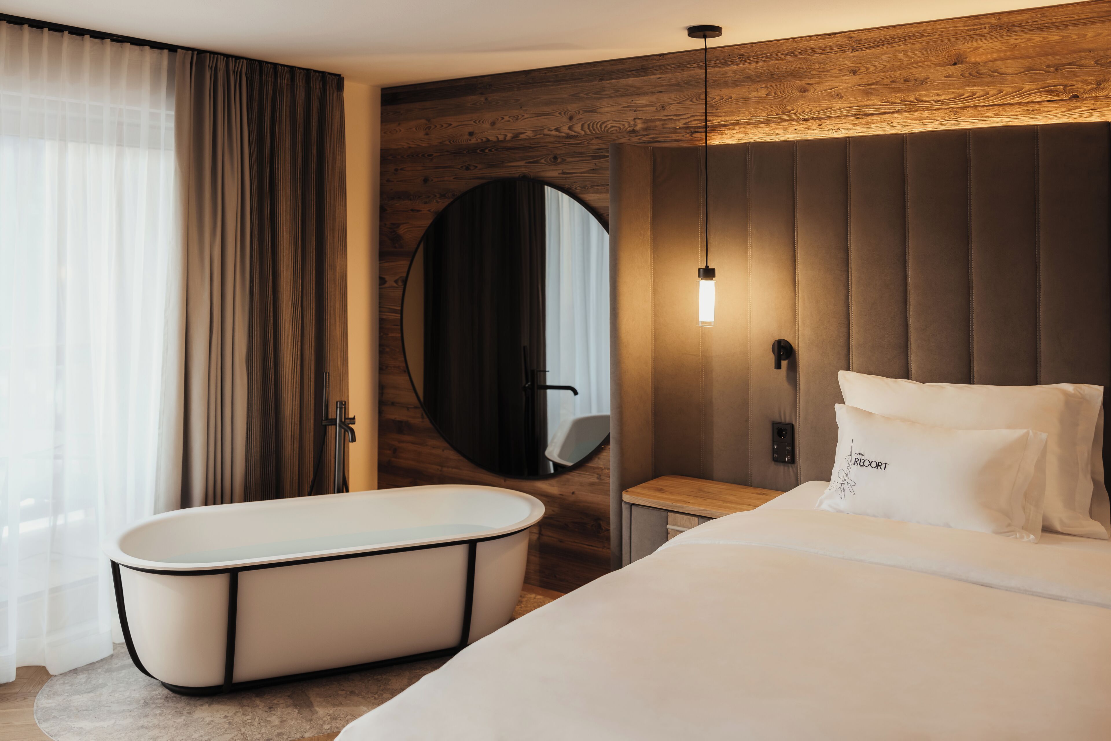 A modern hotel room with a large bed, a bathtub, a wooden wall, and a round mirror.