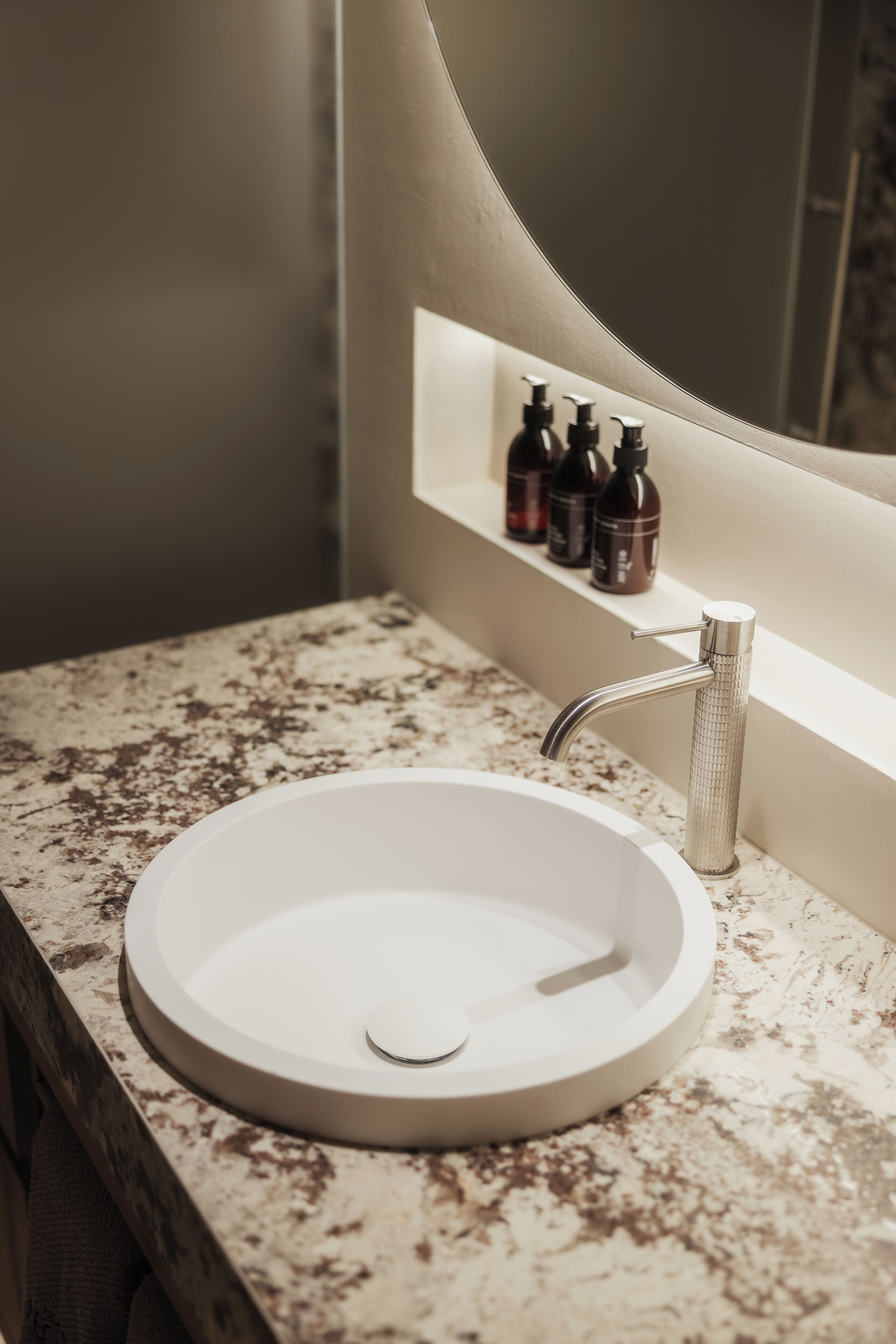 Un baño con un lavabo blanco redondo, un grifo moderno y un estante con tres botellas.