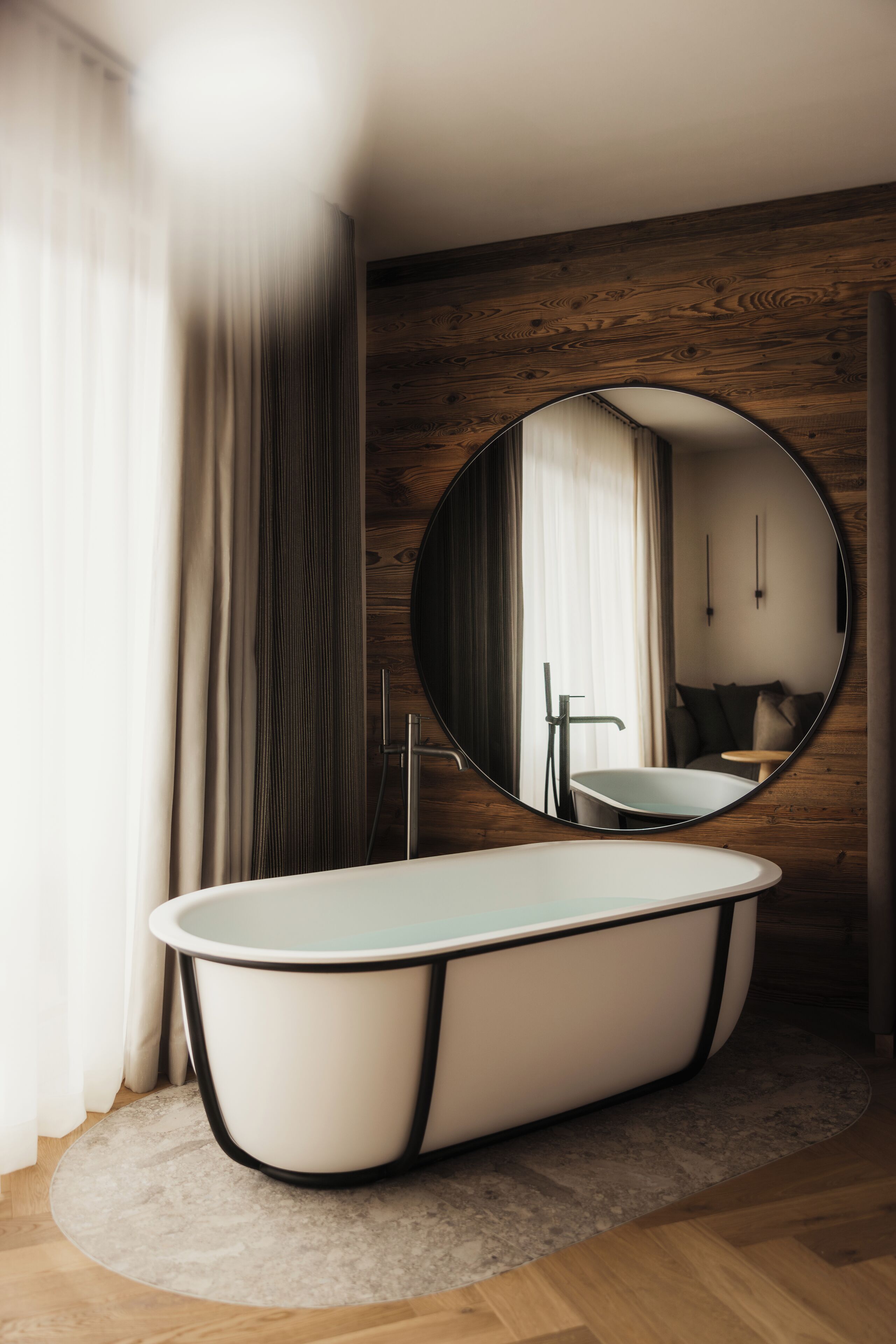 Un baño moderno con una bañera independiente, un espejo redondo, paneles de madera y una ventana con cortinas.