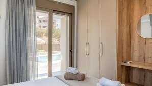 A hotel room with a bed, a wooden wardrobe, a bathroom with a mirror, and a view of a swimming pool. - Villa Leandros With Pool & bbq (Agios Nikolaos)