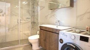 A modern bathroom with a glass-enclosed shower, a toilet, a sink with a wooden cabinet, and a washing machine. - Villa Leandros With Pool & bbq (Agios Nikolaos)