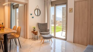 A modern living room with a dining area, a grey armchair, a small side table with a vase, and a large sliding glass door leading to an outdoor area. - Villa Leandros With Pool & bbq (Agios Nikolaos)