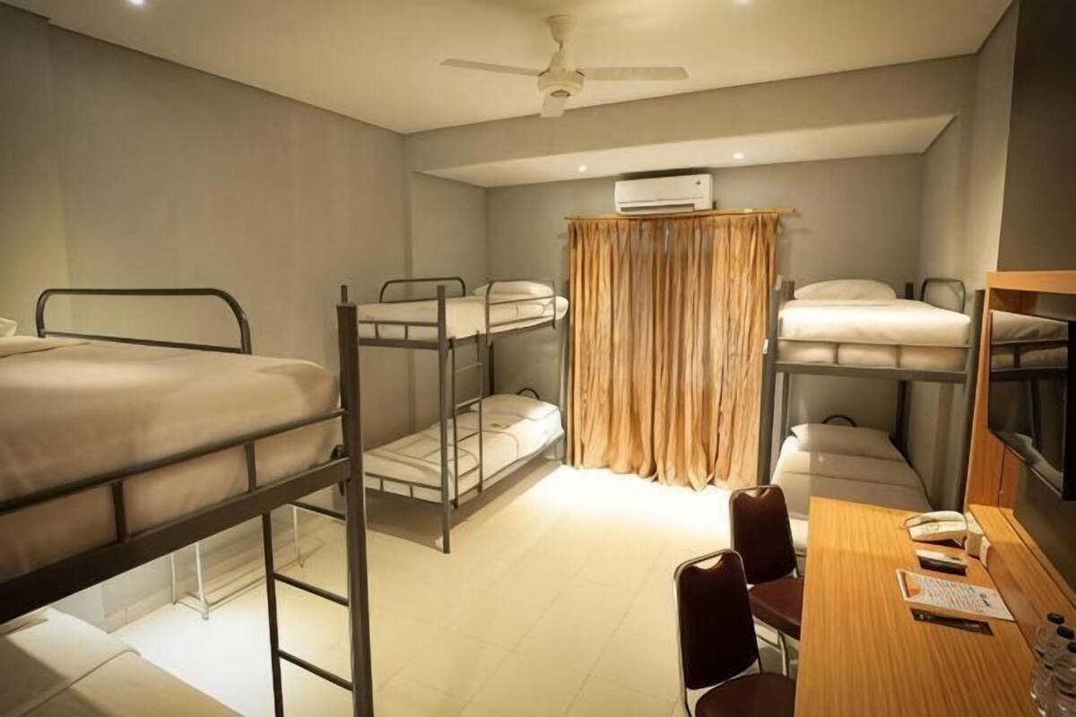 Um quarto de dormitório com beliches, uma escrivaninha, uma cadeira, um ventilador de teto e um ar condicionado.