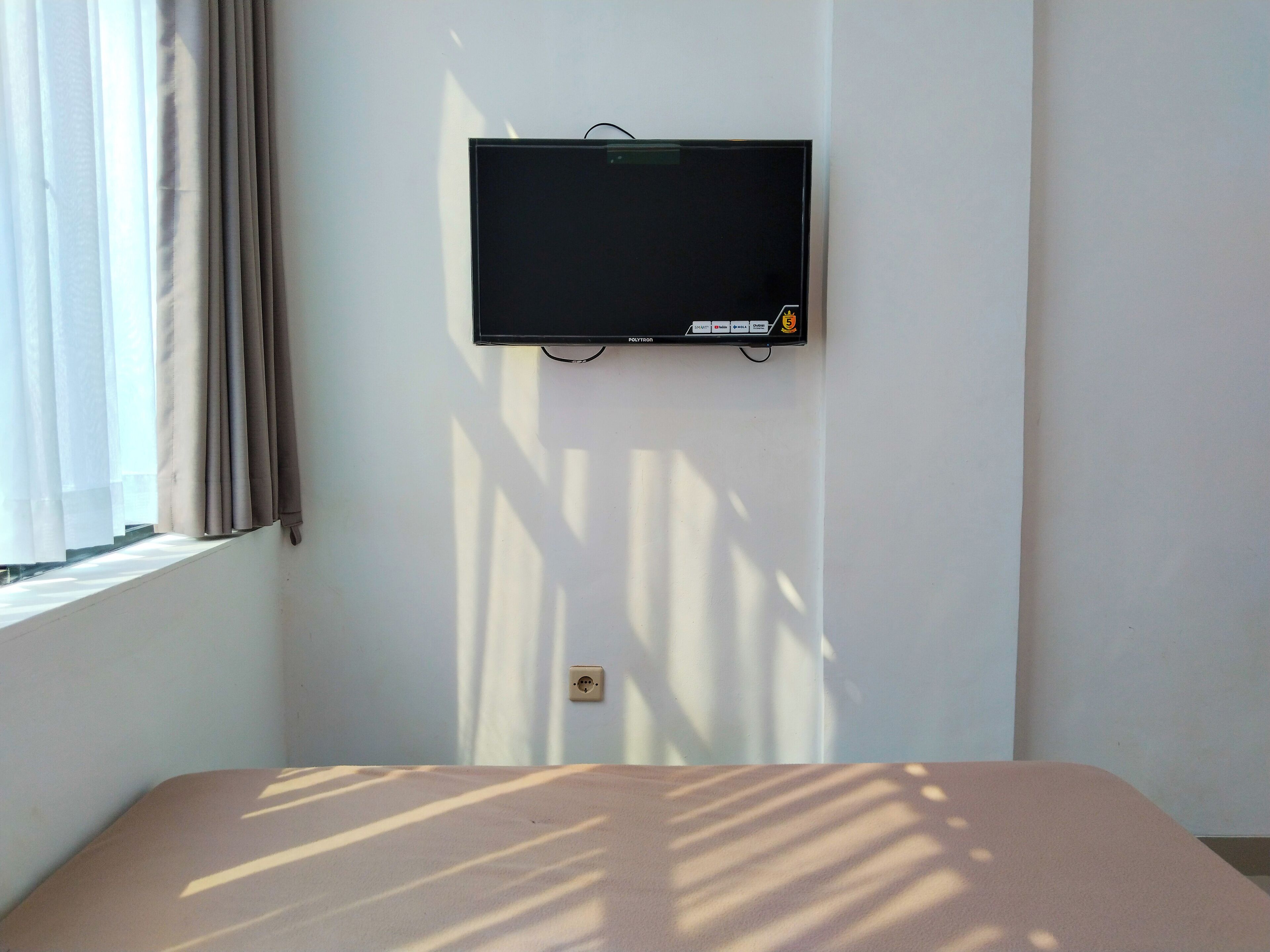 Una habitación con cama, televisor montado en la pared y ventana con cortinas.