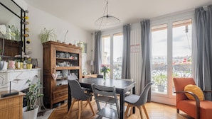 A dining area with a wooden table and chairs, a cabinet with plants, and a view through large windows.