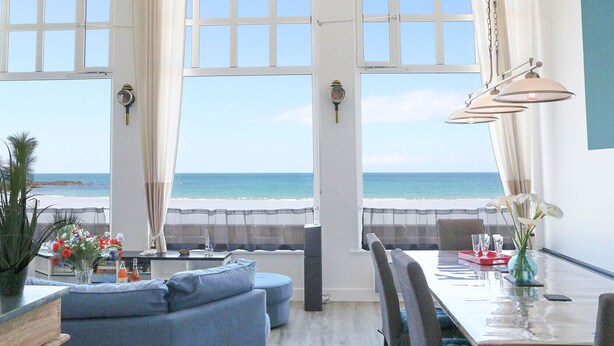 A dining area with a view of the beach, large windows, and a table set with glasses and a vase of flowers. - L'Émeraude Malouine (Saint-Malo)