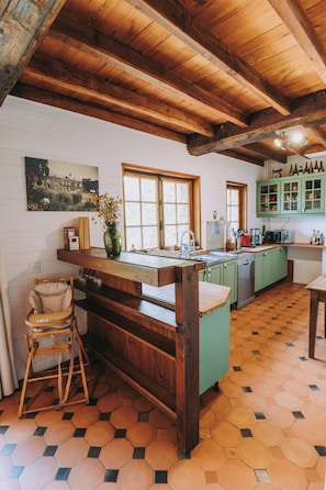 Een keuken met houten balken, een groen kookeiland, een eethoek met een stoel en een zeshoekig betegelde vloer.