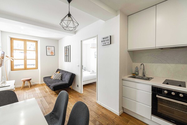 A modern kitchen with white cabinets, a built-in oven, and a sink. There is a black sofa with a geometric pillow, a small wooden stool, and a dining table with chairs.
