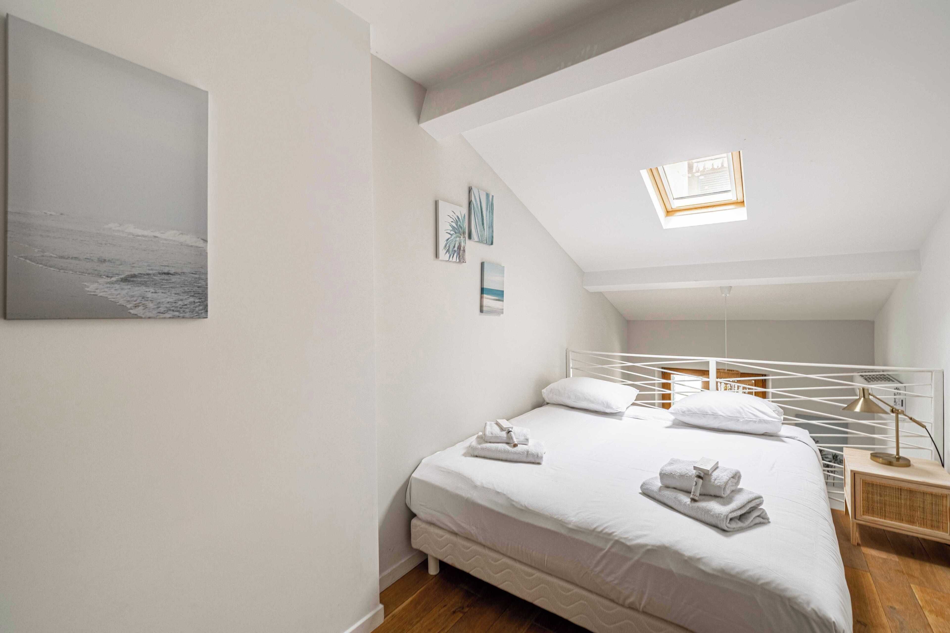 A bedroom with a bed, white walls, a skylight, and a painting of the ocean.