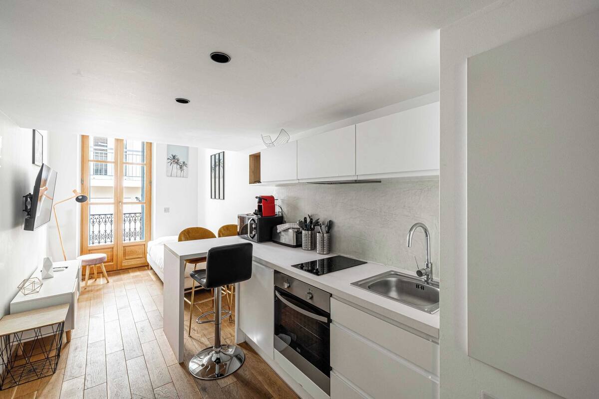 A modern kitchen with white cabinets, a black countertop, and a stainless steel sink. There is a dining area with a white table and chairs, and a small stool.