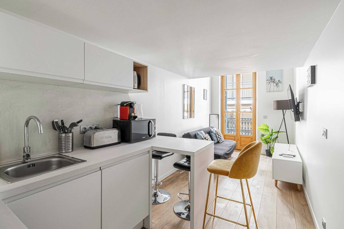 A compact kitchen with a sink, toaster, and microwave. A small bar stool and a plant are near the entrance.