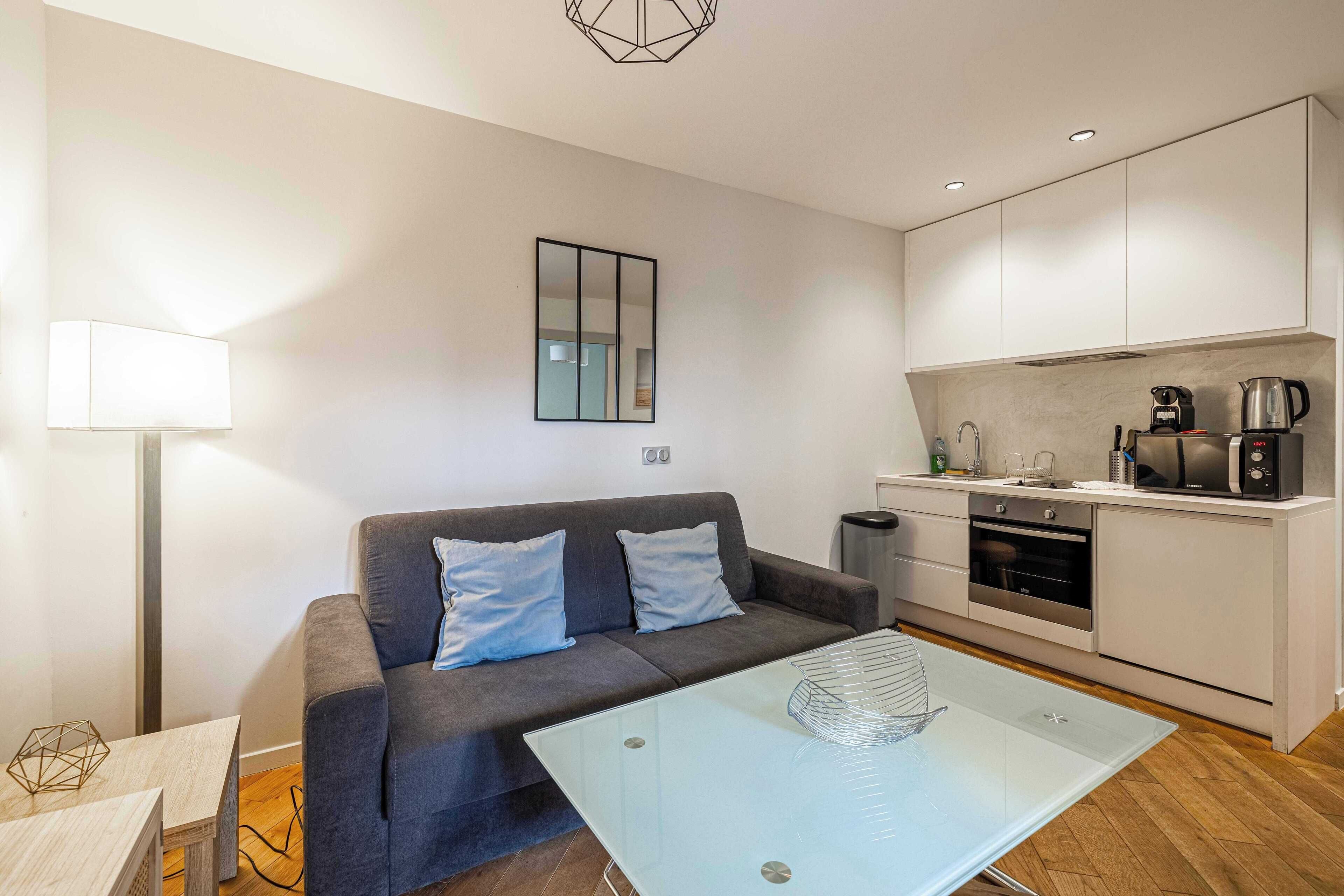 A modern living room with a grey sofa, a glass coffee table, and a kitchen area with white cabinets and appliances.