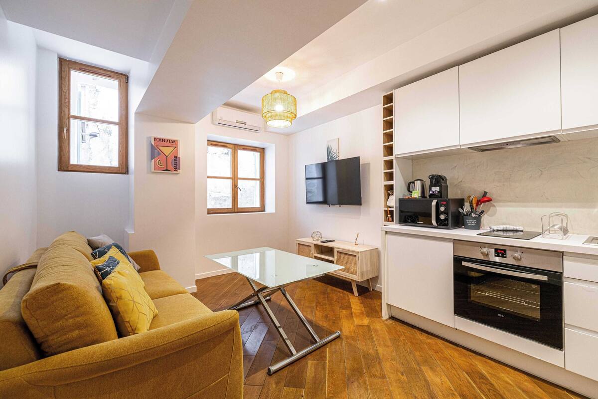 A compact living space with a sofa, a glass-top coffee table, a flat-screen TV, a kitchen with white cabinets, a built-in oven, and a wooden floor.
