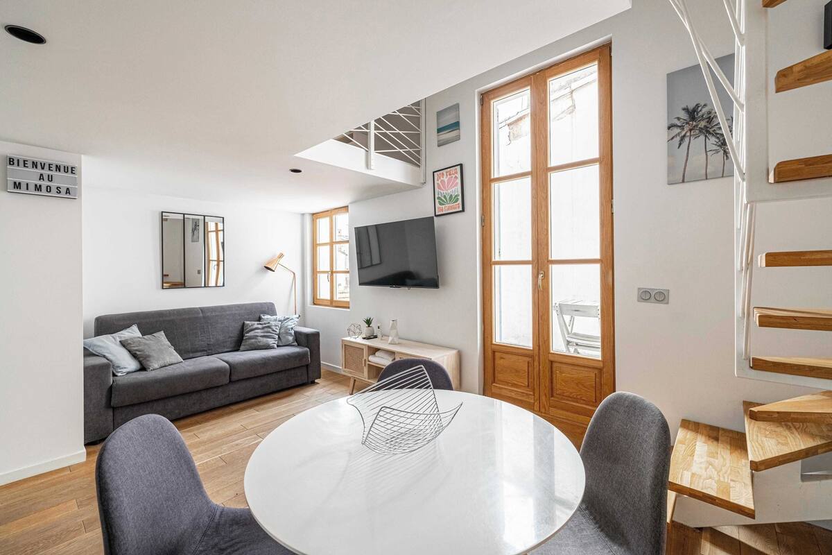 A modern living room with a grey sofa, a round dining table, and a TV mounted on the wall.