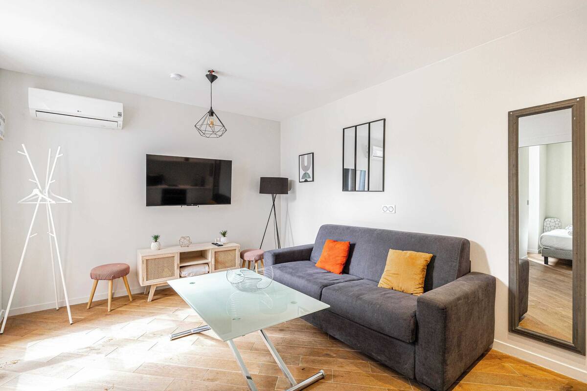 A modern living room with a grey sofa, a glass coffee table, a flat-screen TV mounted on the wall, a standing lamp, and a small side table with a plant.