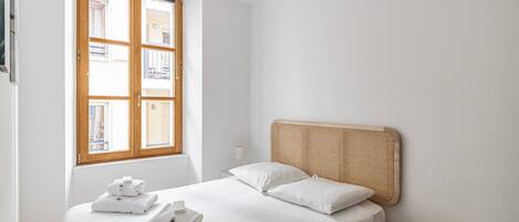 Une chambre à coucher avec un lit, une fenêtre, une table de chevet et un tabouret.