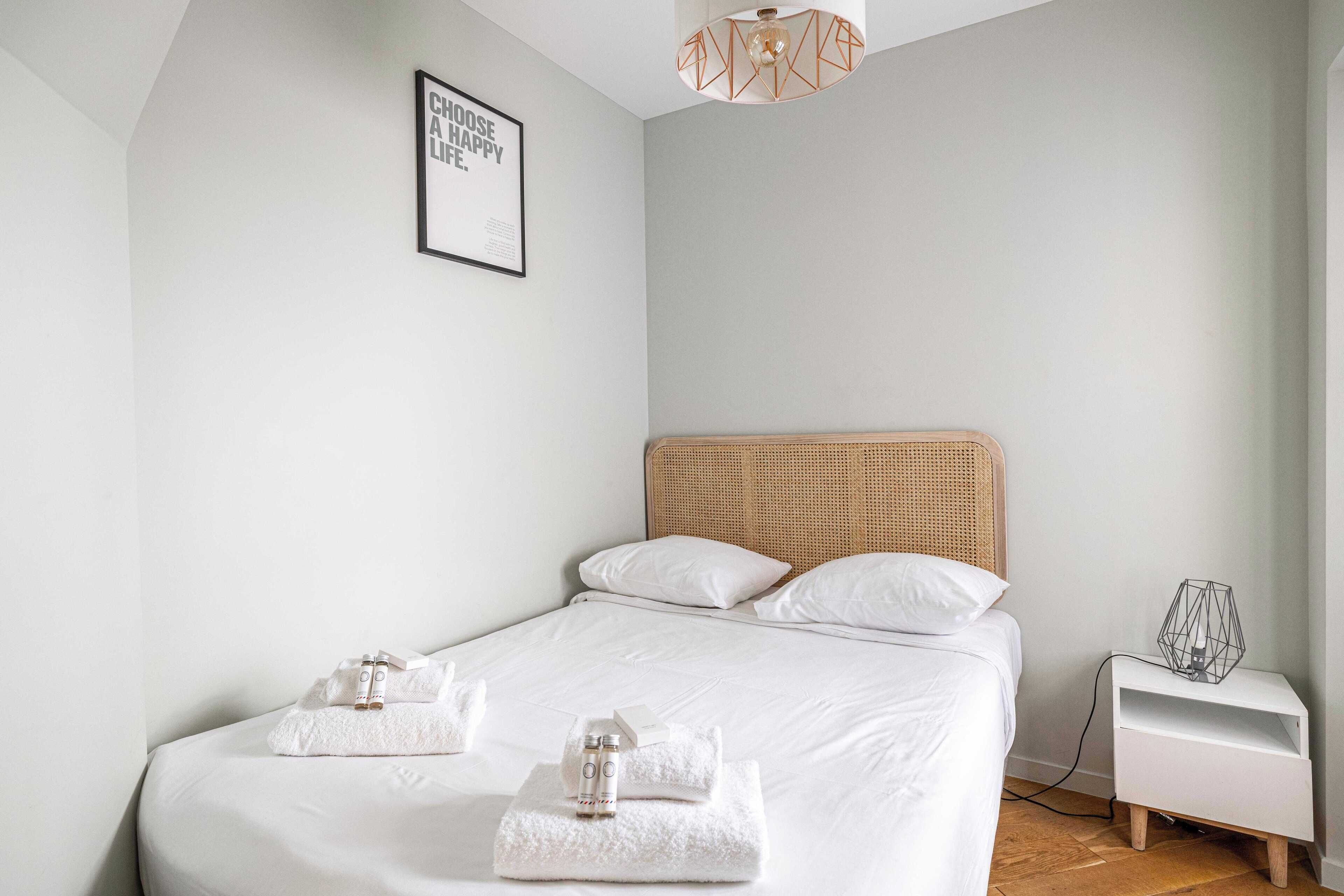 Una cama bien hecha con sábanas blancas, un cabecero de mimbre y una mesita de noche con una lámpara de diseño geométrico.