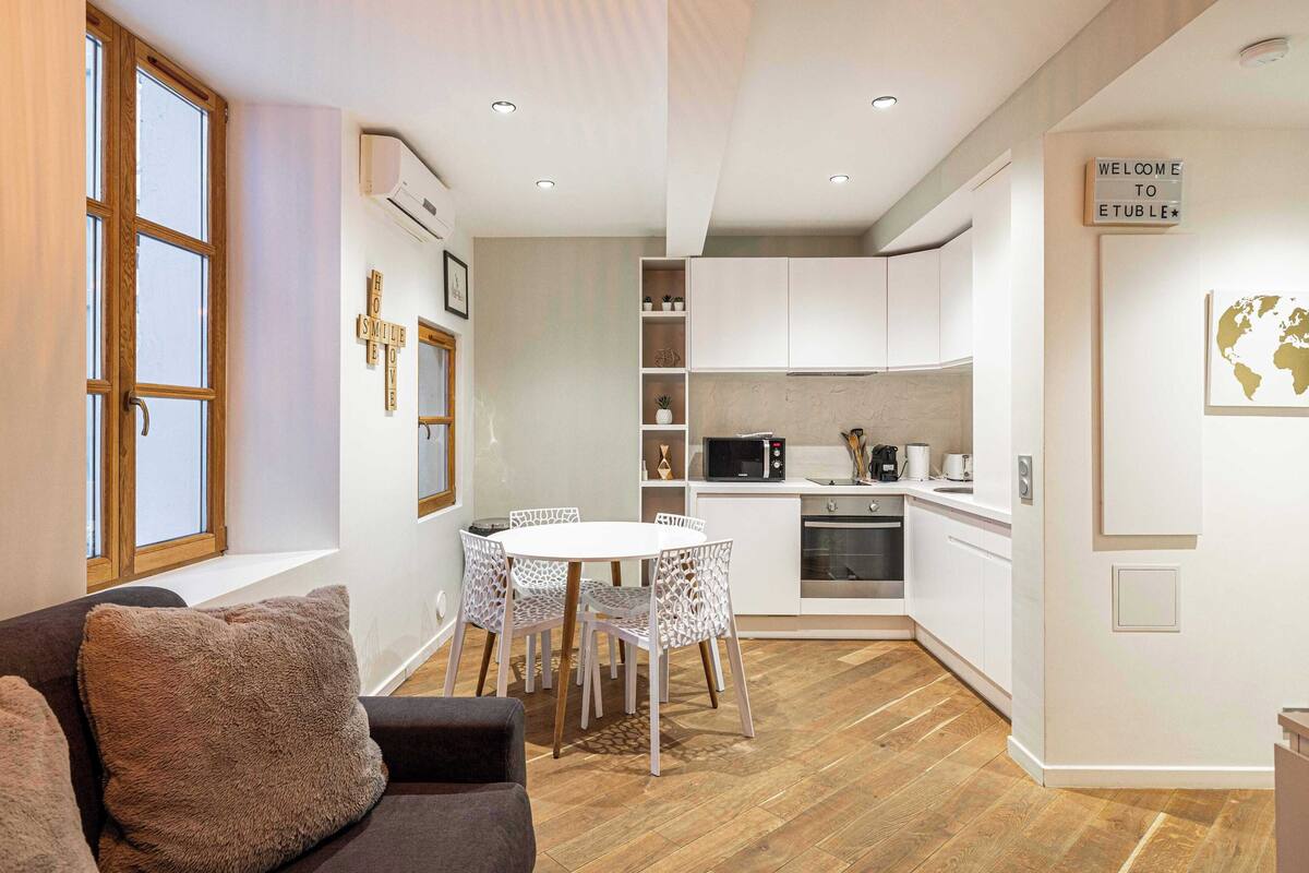 A modern living room with a sofa, a round dining table with chairs, and a kitchen area with white cabinets and appliances.
