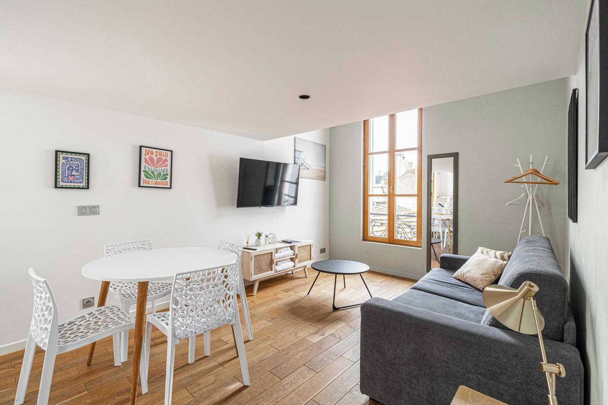A modern living room with a grey sofa, white chairs, a round dining table, and a TV mounted on the wall.
