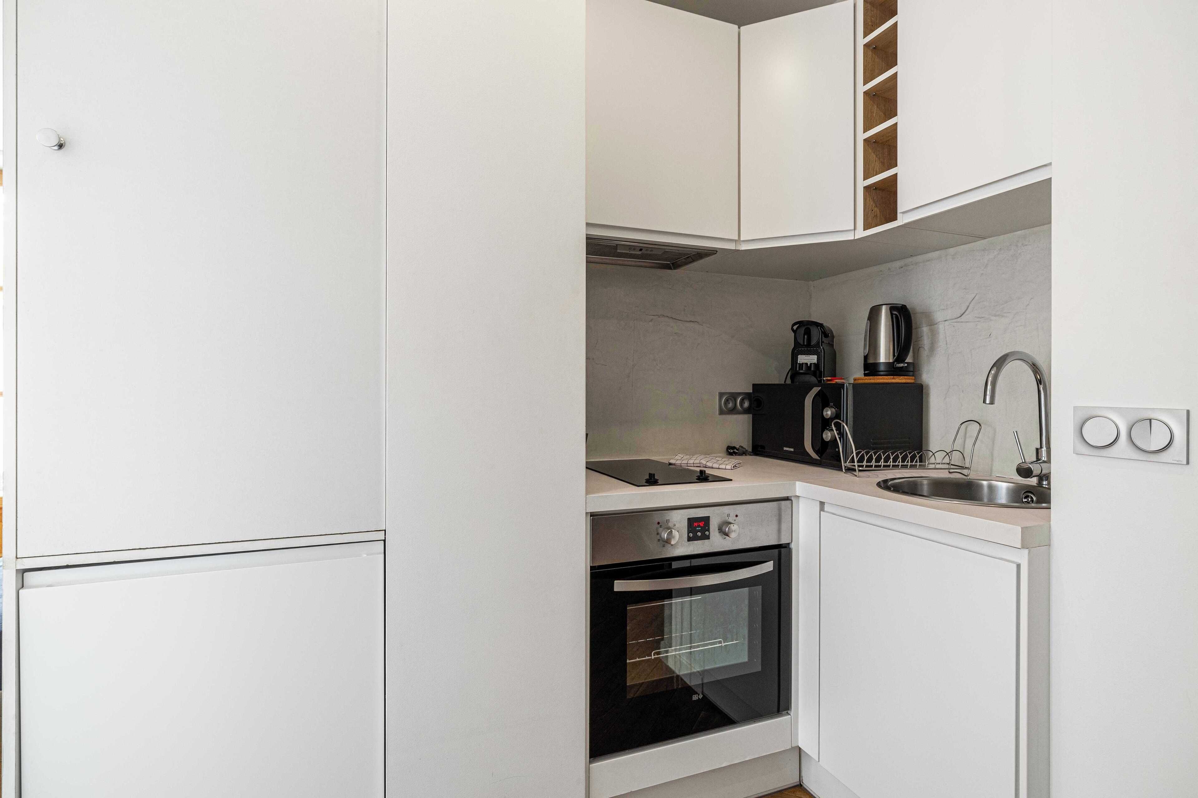 Une cuisine compacte avec des meubles blancs, un four encastré, une machine à café et un évier.