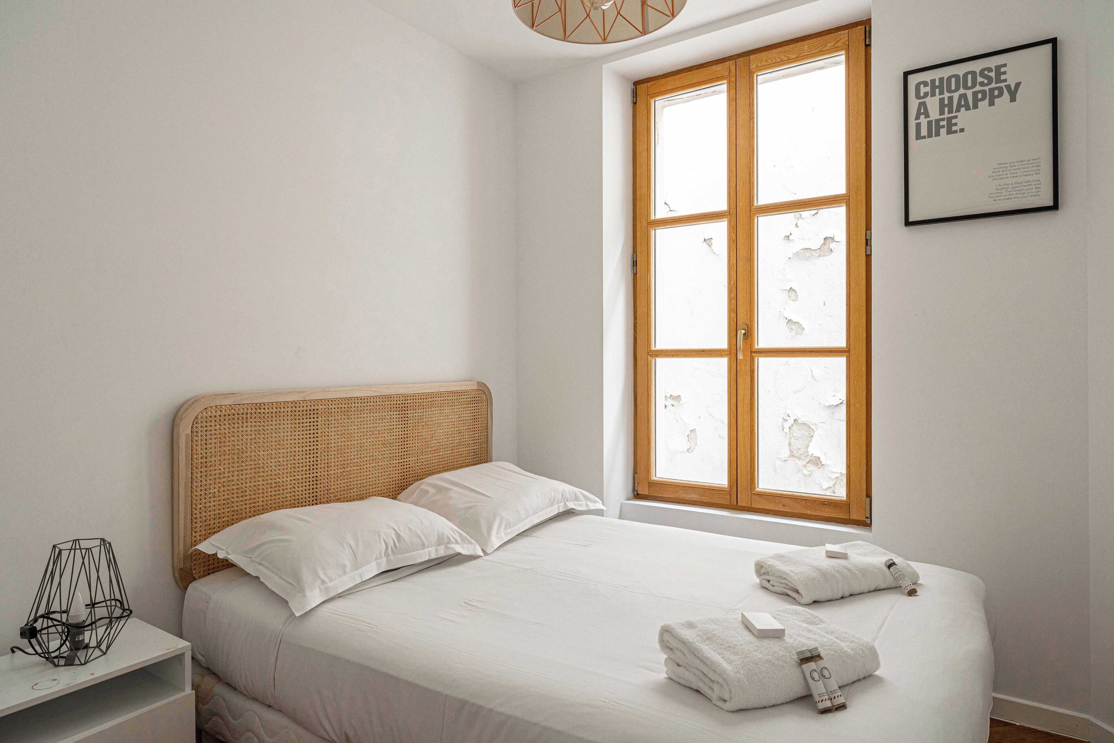 A bedroom with a large window, a bed with white linens, and a framed inspirational quote on the wall.
