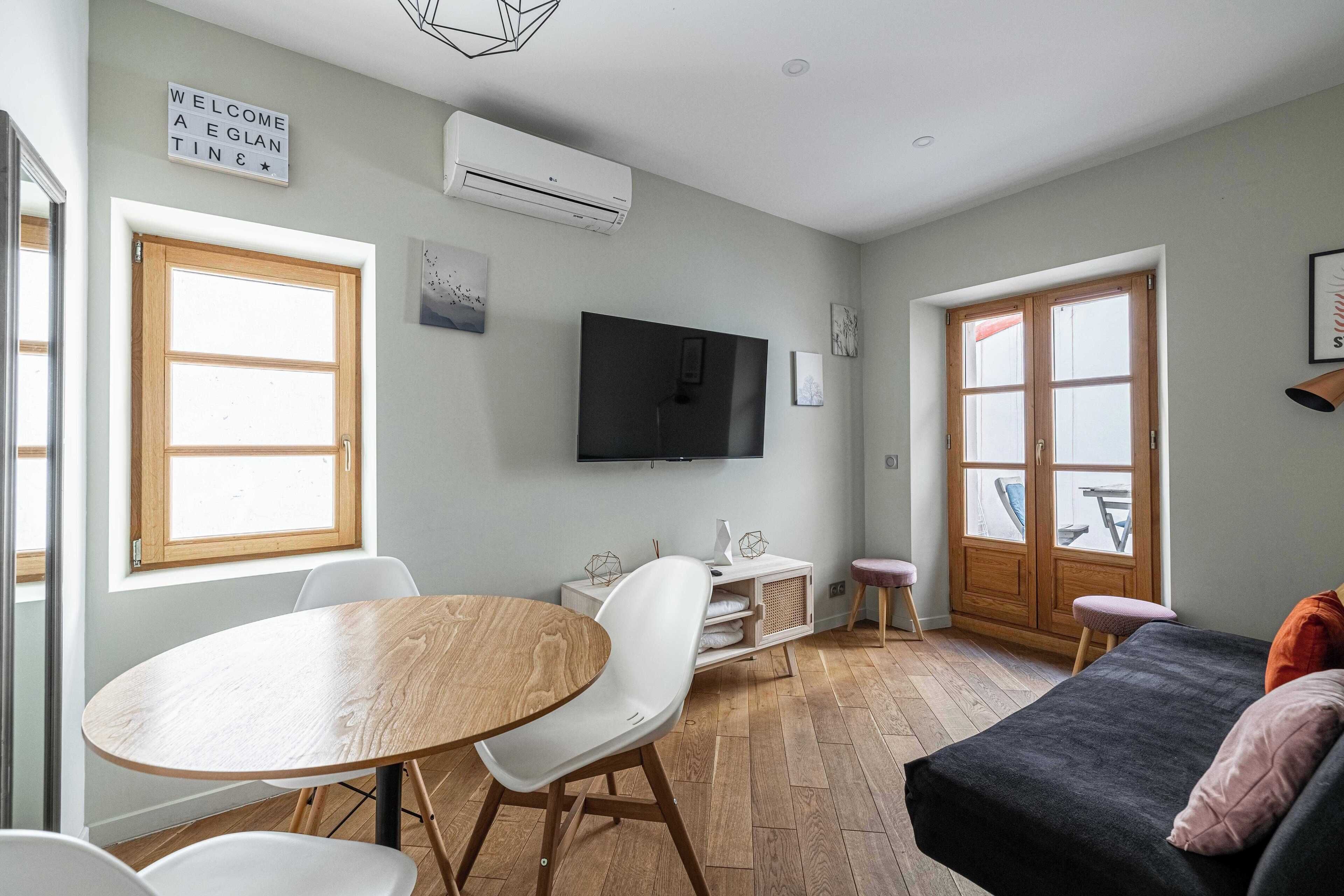 A modern living room with a wooden dining table, white chairs, a black sofa, and a television mounted on the wall.