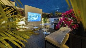 Eine Außenpatio mit einem Fernsehbildschirm, weißen Stühlen, einem Tisch und einem Grill.