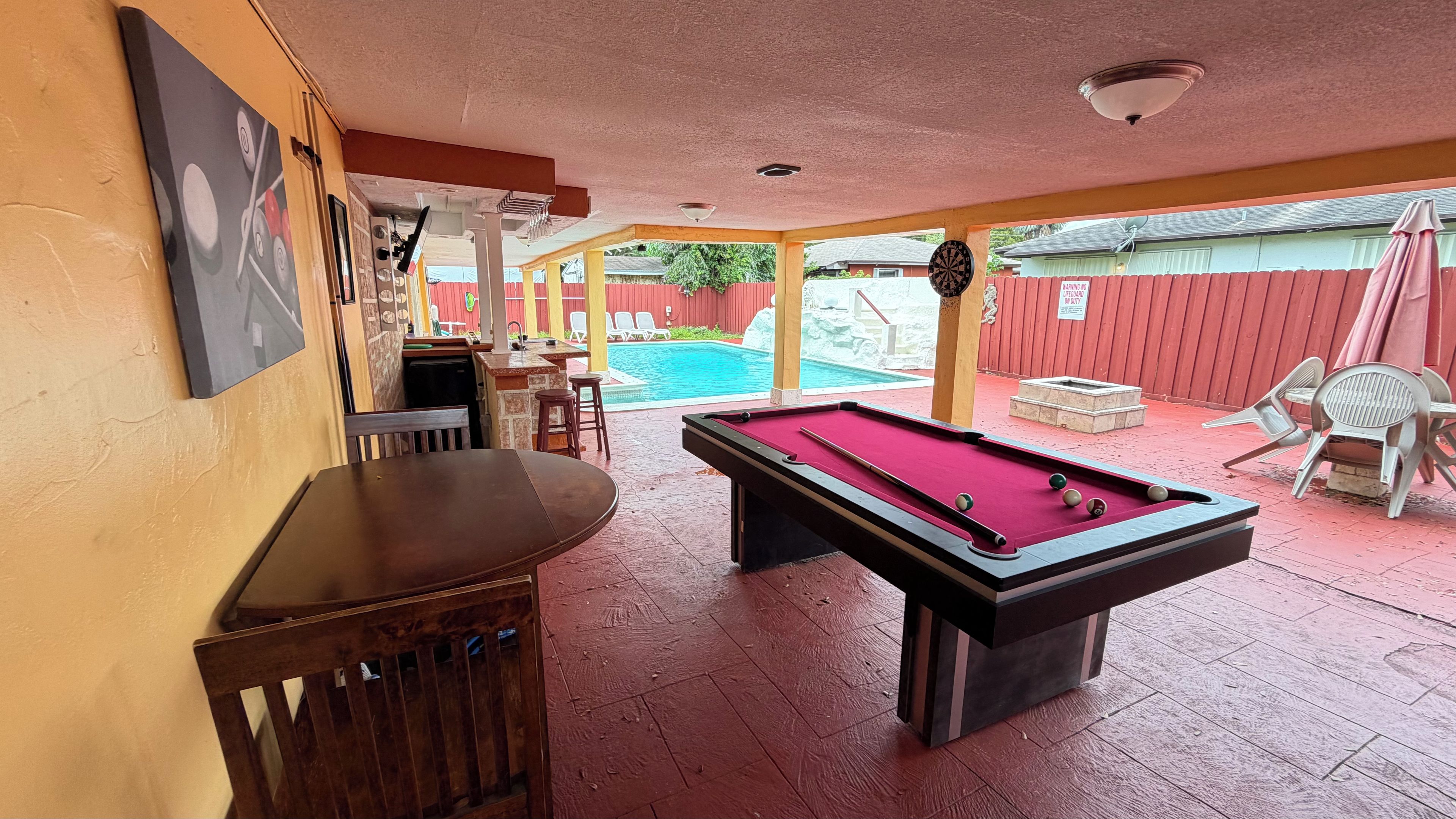 A pool table in a room with a pool, a bar area, and outdoor seating.