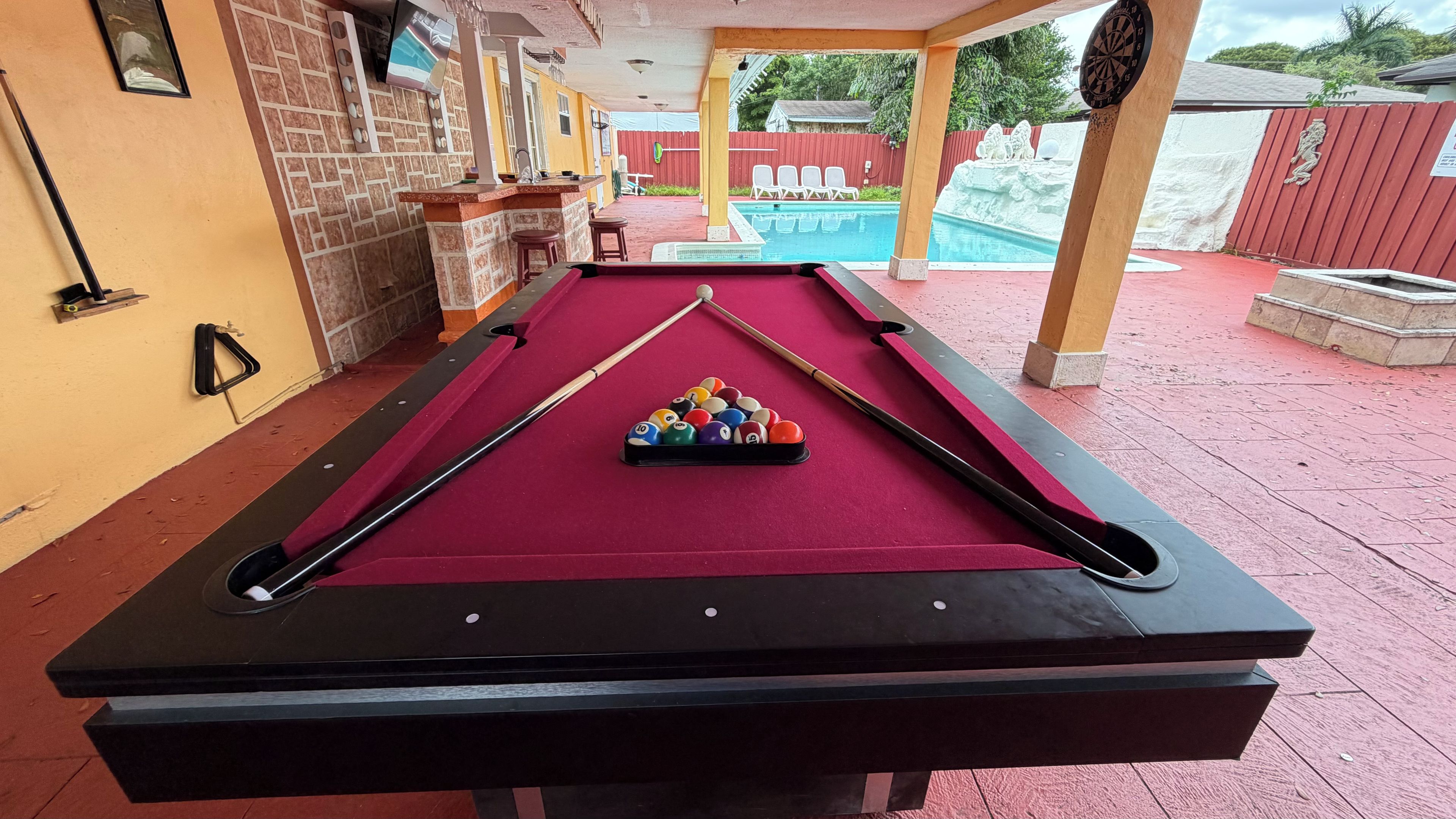 A pool table with a red felt surface, pool cues, and balls, set in a room with a brick wall and a television mounted on the wall.
