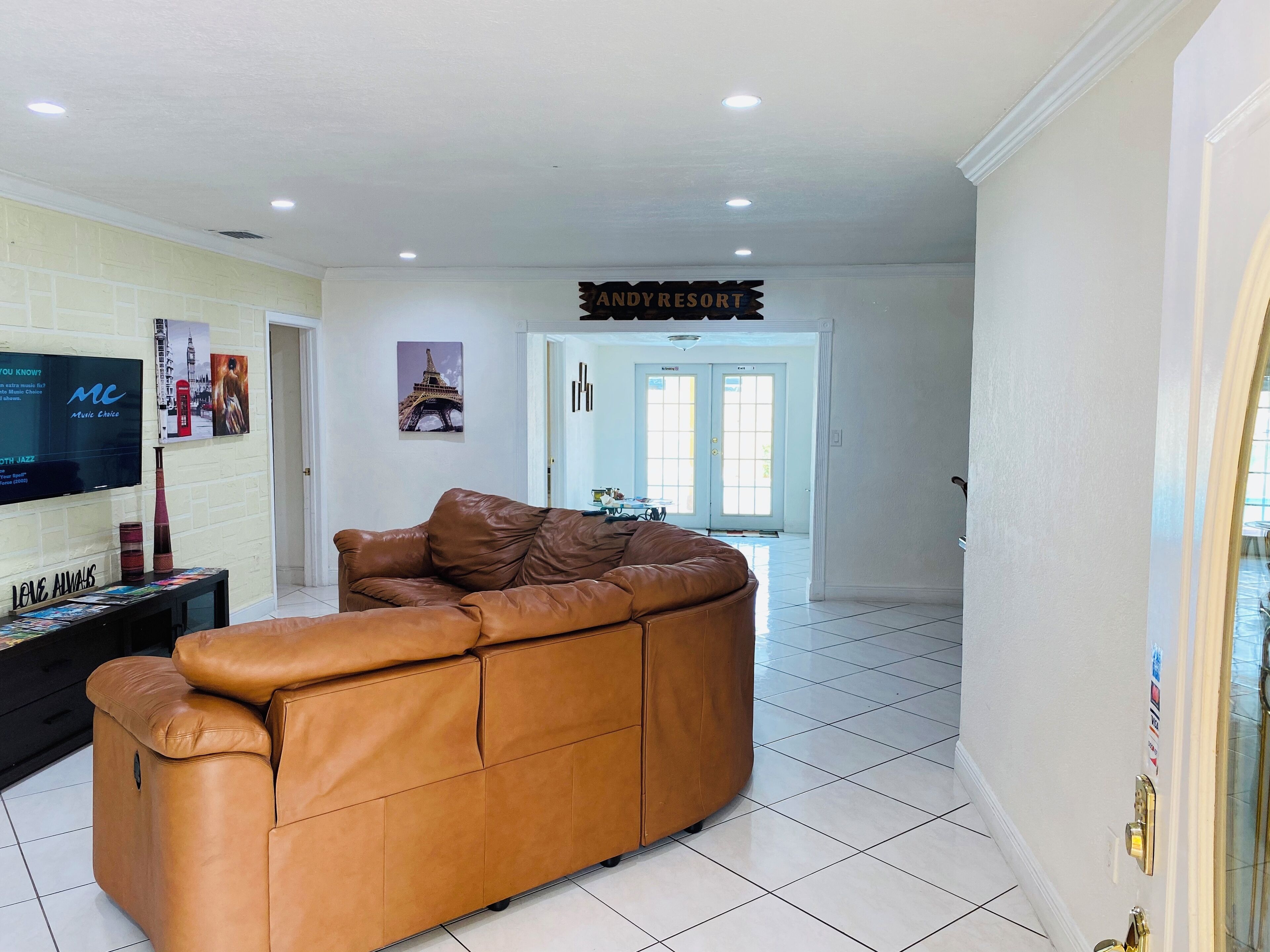 A spacious living room with a large brown leather sofa, a flat-screen TV mounted on the wall, and a sign above the door reading "ANDYRESORT".